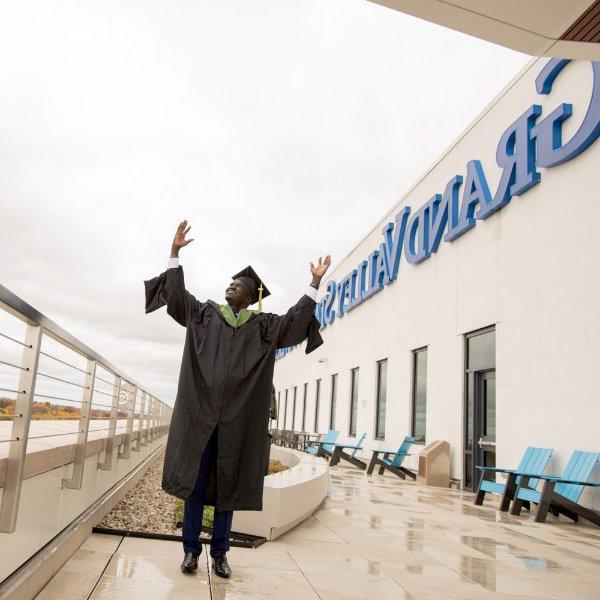 A person wearing a cap and gown has their arms raised. The words Grand Valley State are on a building in the background.